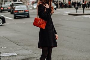 woman in black coat and red leather sling bag standing on sidewalk during daytime