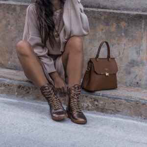 woman sitting on brown concrete surface wearing brown boots beside brown leather handbag