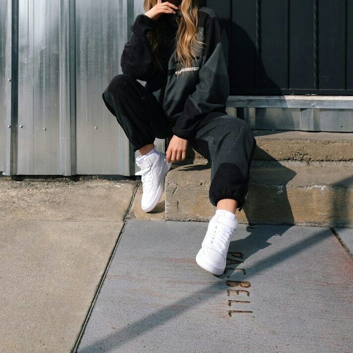 woman in black jacket and white pants sitting on gray concrete floor